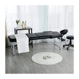 Adjustable height massage table set up in a bright room with white linens and a black cushioned surface.